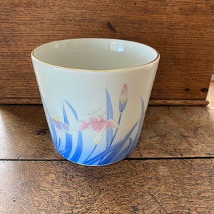 May include: White ceramic cup with a blue and pink floral design. The cup has a gold rim and is decorated with a pattern of irises and leaves.