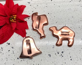 May include: Three copper-colored cookie cutters in the shapes of a bell, a camel, and a saguaro cactus. A red poinsettia flower is in the upper left corner. The cookie cutters are on a speckled white surface.