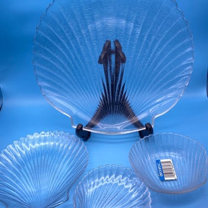 May include: Set of four clear glass serving dishes shaped like seashells. The largest dish is on a stand and the smaller dishes are on a blue surface. The smaller dishes have a sticker that says "arcopal".