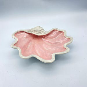 May include: A ceramic seashell-shaped bowl with a wavy, scalloped edge. The interior is a swirl of pink and white, while the rim and exterior are a creamy white. This decorative bowl is ideal for serving snacks or as a decorative accent.