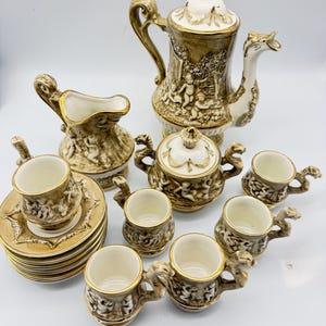 May include: A ceramic tea set featuring a coffee pot, a lidded sugar bowl, a creamer, and six cups with saucers. Each piece is finished in a metallic gold and white glaze, decorated with embossed cherub figures and ornate, curved handles.