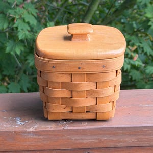 May include: A square woven wooden basket with a light brown finish and a matching wooden lid. The lid features a small, centered square handle. The basket body is constructed from horizontal and vertical wooden strips secured with small metal nails.