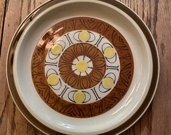May include: A large, round ceramic plate with a brown and white geometric design. The plate has a brown rim and a white center with a brown and yellow pattern.