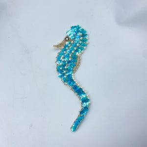 May include: A sequined seahorse brooch in shades of turquoise and gold. The seahorse is embellished with turquoise sequins and small white beads, with a gold-colored snout and a delicate, curved shape. The brooch is likely for clothing or accessories.