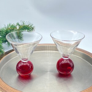 May include: Two clear martini glasses with a red, spherical base sit on a silver tray with a copper-colored rim. The glasses have a classic cone-shaped bowl. A sprig of green pine needles is in the background.