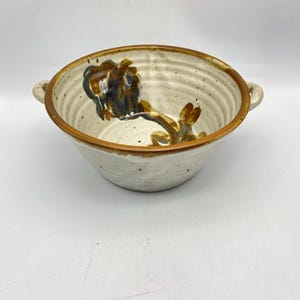 May include: A white ceramic bowl with brown and blue painted floral designs. The bowl has two handles and a slightly rounded rim.