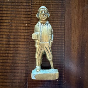 May include: A vintage brass figurine of a man in a hat and coat, holding a small object. The figure has a smiling face and is standing on a rectangular base. The brass has a slightly aged patina.
