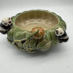 Puede incluir: Un cuenco de cerámica con exterior verde en forma de calabaza, con un pequeño tallo y hojas de calabaza. Dos figuras de oso panda están posadas en los lados. El interior es de color beige claro.