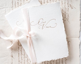 May include: Two white paper cards with a pink ribbon tied around them. The top card has the words "Our Vows" written in cursive.