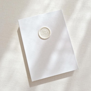 May include: A white rectangular card with a circular wax seal that reads "OUR VOWS" in a serif font. The card is centered on a white textured surface, with shadows from overhead.