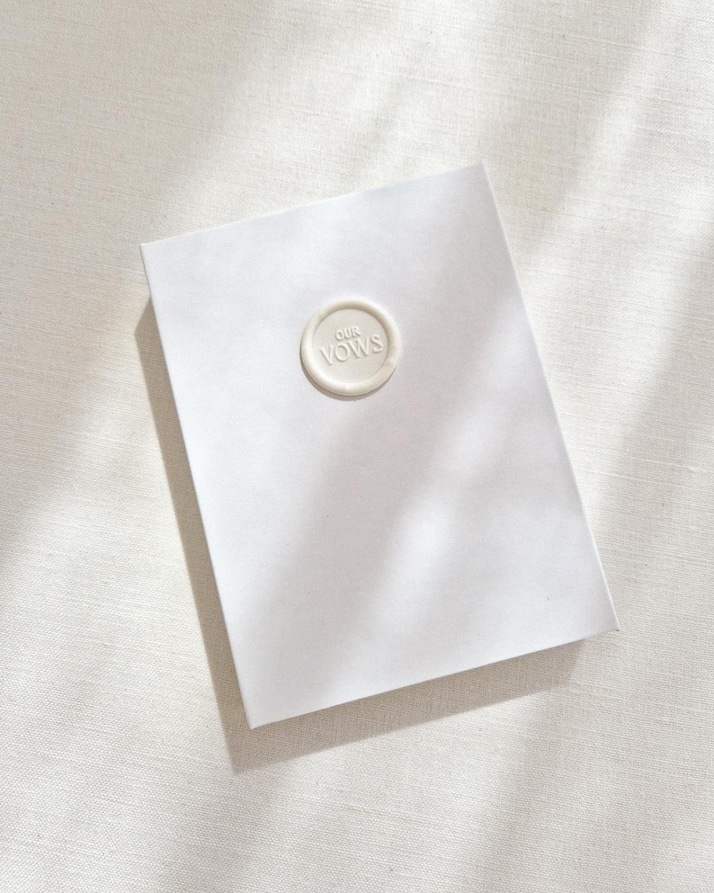 May include: A white rectangular card with a circular wax seal that reads "OUR VOWS" in a serif font. The card is centered on a white textured surface, with shadows from overhead.