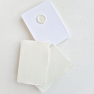 May include: A white stationery set featuring a sealed envelope and two small cards. The envelope is sealed with a circular wax stamp. The cards are embossed with the words "Her Vows" and "His".
