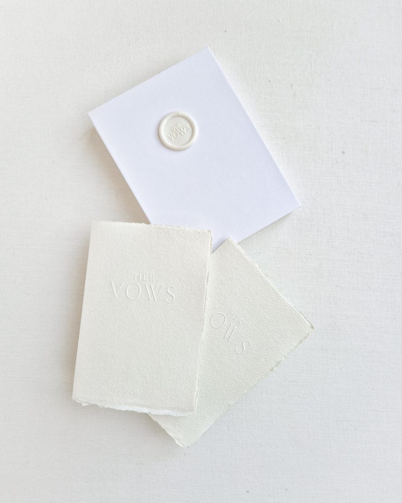 May include: A white stationery set featuring a sealed envelope and two small cards. The envelope is sealed with a circular wax stamp. The cards are embossed with the words "Her Vows" and "His".