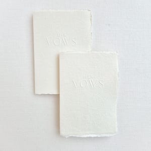 May include: Two off-white vow books with deckled edges. The top book reads "HIS VOWS" in embossed lettering. The bottom book reads "HER VOWS" in embossed lettering. The books are rectangular and sit on a white surface.