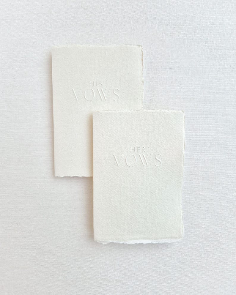May include: Two off-white vow books with deckled edges. The top book reads "HIS VOWS" in embossed lettering. The bottom book reads "HER VOWS" in embossed lettering. The books are rectangular and sit on a white surface.