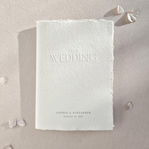 Könnte beinhalten: Ein rechteckiges, cremeweißes Hochzeitsgästebuch mit einem strukturierten Papierumschlag mit Büttenrand. Der Umschlag zeigt den geprägten Text "OUR WEDDING" und den schwarzen Aufdruck "SOPHIA & ALEXANDER" sowie das Datum "AUGUST 21, 2027" unten.