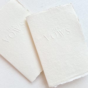 May include: Two off-white vow books with deckled edges. The words "HIS VOWS" and "HER VOWS" are embossed on the covers. The books are rectangular and made of textured paper, likely for a wedding or vow renewal.