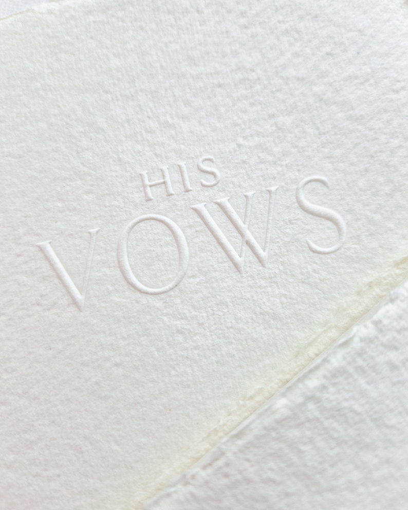 May include: Close-up of a textured, off-white paper with the embossed words "HIS VOWS". The paper has a deckled edge and a subtle, natural appearance, perfect for wedding stationery.