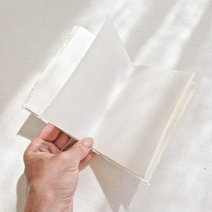 May include: An open, off-white journal with blank pages. The journal has a textured, fringed edge. The pages are held open by a hand with a gold bracelet. The journal is on a white surface.