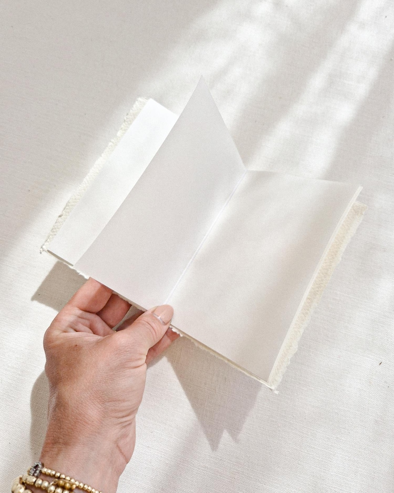 May include: An open, off-white journal with blank pages. The journal has a textured, fringed edge. The pages are held open by a hand with a gold bracelet. The journal is on a white surface.