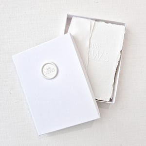 May include: A white box with an open lid, revealing textured white paper. The box has a circular seal that reads "OUR VOWS". The paper inside is embossed with the words "OUR VOWS". The overall theme is wedding stationery.
