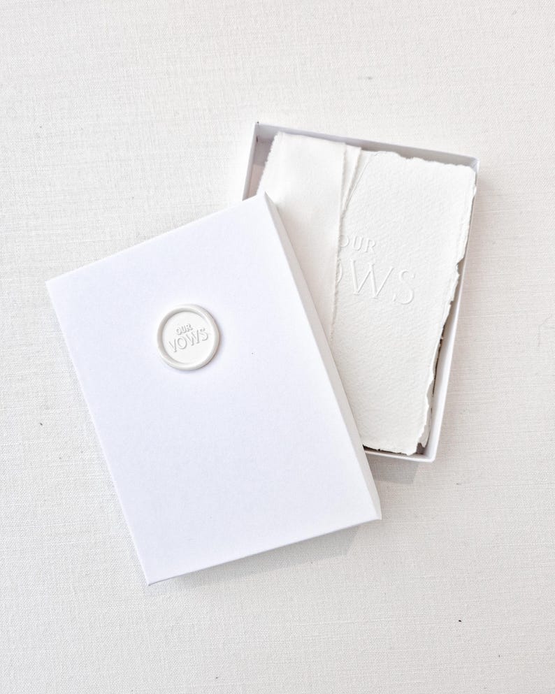 May include: A white box with an open lid, revealing textured white paper. The box has a circular seal that reads "OUR VOWS". The paper inside is embossed with the words "OUR VOWS". The overall theme is wedding stationery.