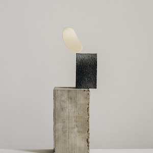 Op de afbeelding: Een minimalistisch stilleven met een lichtbruine ovale vorm die in evenwicht is op een donkergrijze rechthoekige blok. De blok staat op een grotere, lichtgrijze rechthoekige blok.
