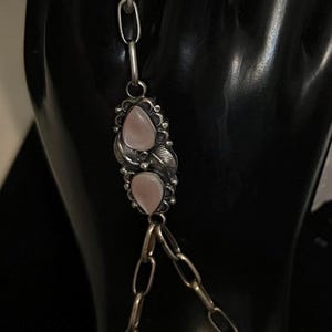 Pode incluir: Uma corrente de m&atilde;o de prata com um anel, corrente e pulseira. A joia apresenta pedras rosa em forma de p&ecirc;ra e detalhes de folhas. O anel e a pulseira s&atilde;o conectados por uma corrente com duas pedras rosa adicionais.