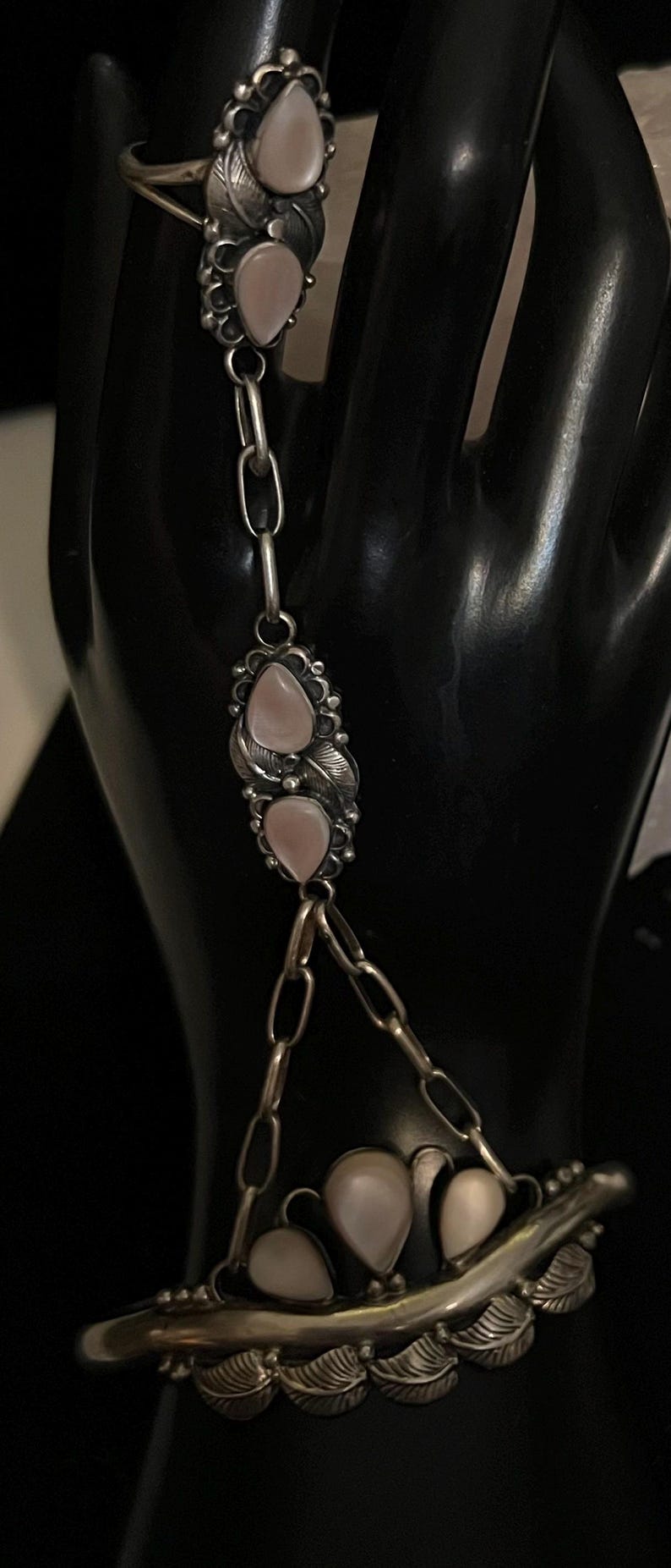 Pode incluir: Uma corrente de m&atilde;o de prata com um anel, corrente e pulseira. A joia apresenta pedras rosa em forma de p&ecirc;ra e detalhes de folhas. O anel e a pulseira s&atilde;o conectados por uma corrente com duas pedras rosa adicionais.