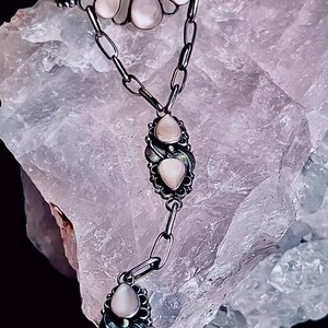 Pode incluir: Um conjunto de colar e anel de prata com pedras preciosas rosa claro em forma de p&ecirc;ra. O colar tem um design de corrente com um pingente decorativo. O anel tem um design semelhante. A joia &eacute; colocada sobre um fundo texturizado roxo claro.