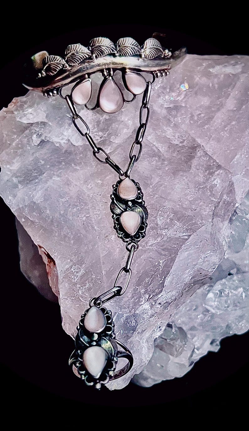 Pode incluir: Um conjunto de colar e anel de prata com pedras preciosas rosa claro em forma de p&ecirc;ra. O colar tem um design de corrente com um pingente decorativo. O anel tem um design semelhante. A joia &eacute; colocada sobre um fundo texturizado roxo claro.