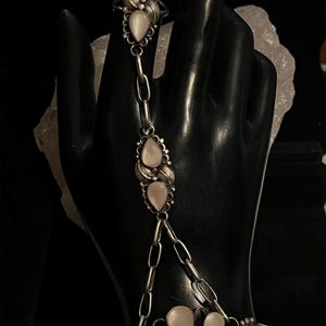Pode incluir: Uma pulseira de corrente de m&atilde;o prateada com pedras peroladas e um anel. O anel e a pulseira s&atilde;o conectados por uma corrente com pedras peroladas adicionais. A pulseira tem um design de folhas.