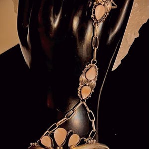 Pode incluir: Uma pulseira de corrente de m&atilde;o prateada com pedras preciosas rosa claro em forma de p&ecirc;ra. A pulseira conecta um anel a uma pulseira de pulso com uma corrente. A pulseira tem detalhes de folhas e tr&ecirc;s pedras preciosas. O anel tem duas pedras preciosas.