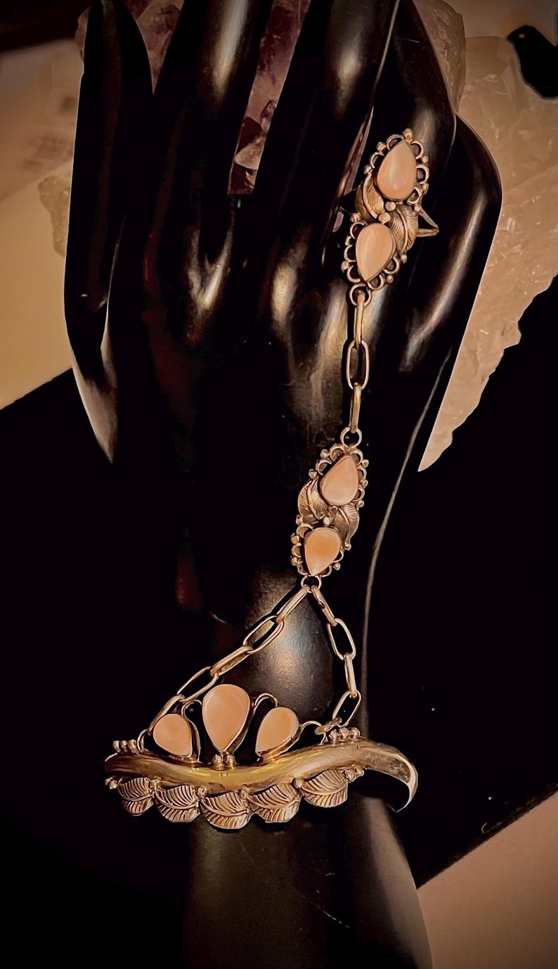 Pode incluir: Uma pulseira de corrente de m&atilde;o prateada com pedras preciosas rosa claro em forma de p&ecirc;ra. A pulseira conecta um anel a uma pulseira de pulso com uma corrente. A pulseira tem detalhes de folhas e tr&ecirc;s pedras preciosas. O anel tem duas pedras preciosas.