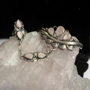 Pode incluir: Um conjunto de anel e pulseira prateados com pedras peroladas em forma de l&aacute;grima. O anel apresenta duas pedras e um design de folha, ligado por uma corrente a uma pulseira com v&aacute;rias pedras e detalhes de folha. As joias est&atilde;o sobre um fundo claro e texturizado.