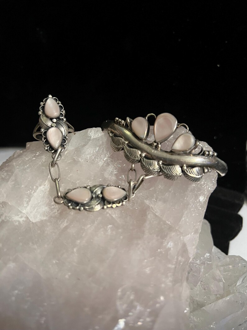 Pode incluir: Um conjunto de anel e pulseira prateados com pedras peroladas em forma de l&aacute;grima. O anel apresenta duas pedras e um design de folha, ligado por uma corrente a uma pulseira com v&aacute;rias pedras e detalhes de folha. As joias est&atilde;o sobre um fundo claro e texturizado.
