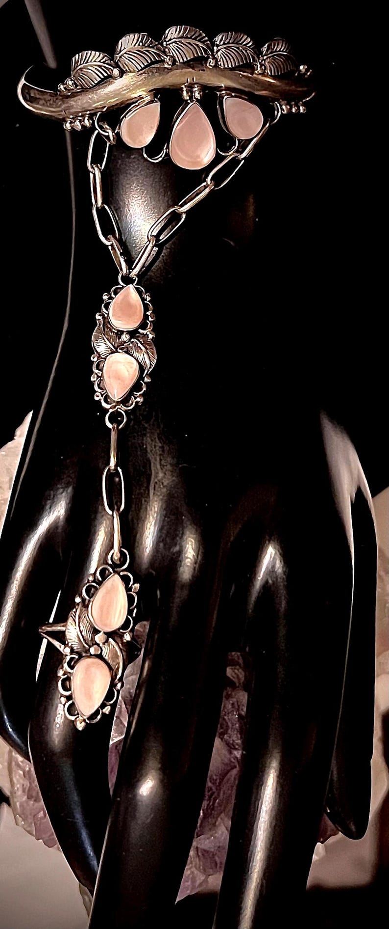 Pode incluir: Uma pulseira de corrente de m&atilde;o prateada com pedras rosa em forma de gota e detalhes de folhas. A pulseira apresenta uma corrente que se conecta a um anel. A joia &eacute; exibida em um fundo preto.