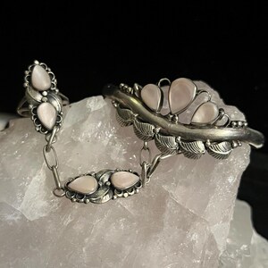 Pode incluir: Um conjunto de anel e pulseira prateados, ligados por uma corrente. As joias apresentam pedras rosa claro em forma de p&ecirc;ra e detalhes em forma de folha. A pulseira tem quatro pedras, e o anel tem duas.