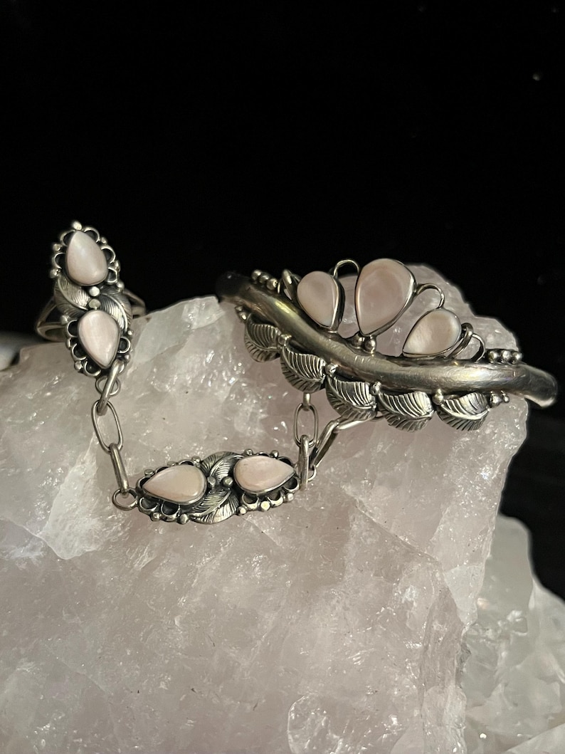 Pode incluir: Um conjunto de anel e pulseira prateados, ligados por uma corrente. As joias apresentam pedras rosa claro em forma de p&ecirc;ra e detalhes em forma de folha. A pulseira tem quatro pedras, e o anel tem duas.