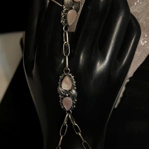 Pode incluir: Uma pulseira de corrente de m&atilde;o prateada com um anel, corrente e componentes de pulseira. A joia apresenta pedras rosa claro em forma de p&ecirc;ra e detalhes de folhas. O anel e a pulseira s&atilde;o conectados por uma corrente com duas pedras rosa adicionais.