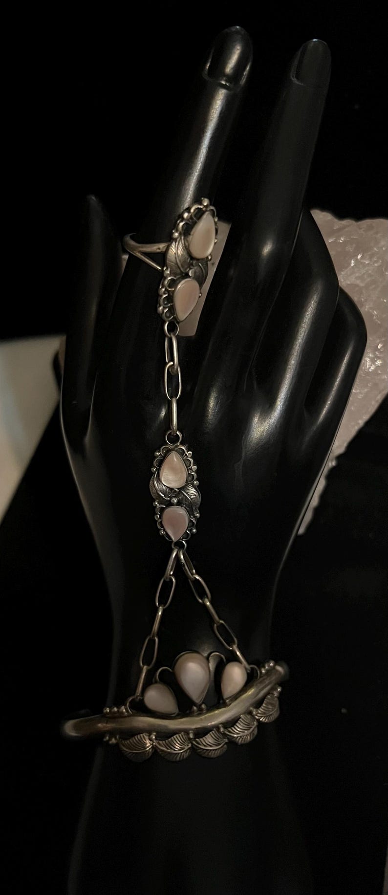 Pode incluir: Uma pulseira de corrente de m&atilde;o prateada com um anel, corrente e componentes de pulseira. A joia apresenta pedras rosa claro em forma de p&ecirc;ra e detalhes de folhas. O anel e a pulseira s&atilde;o conectados por uma corrente com duas pedras rosa adicionais.