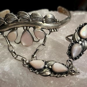 Pode incluir: Um conjunto de joias prateadas com uma pulseira de punho, um anel e uma corrente. A pulseira e o anel s&atilde;o adornados com desenhos de folhas e pedras preciosas rosa claro em forma de gota. O anel &eacute; conectado &agrave; pulseira por uma corrente.