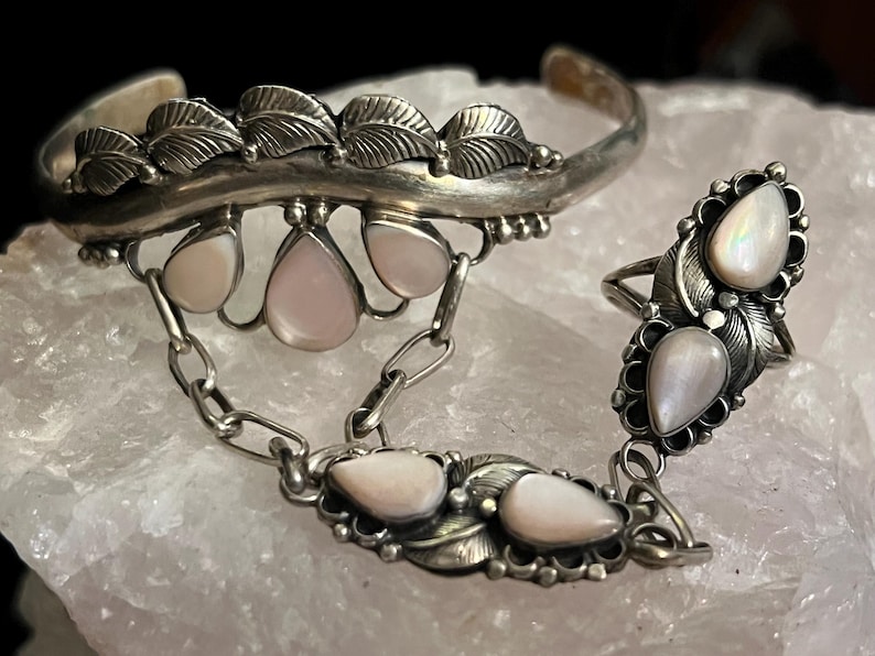 Pode incluir: Um conjunto de joias prateadas com uma pulseira de punho, um anel e uma corrente. A pulseira e o anel s&atilde;o adornados com desenhos de folhas e pedras preciosas rosa claro em forma de gota. O anel &eacute; conectado &agrave; pulseira por uma corrente.