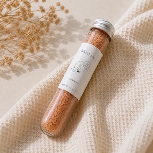 May include: A clear glass tube filled with pink bath salts, labeled "BATH SALT" with a grapefruit illustration. The tube is capped with a silver lid and rests on a cream-colored, textured fabric. Net weight: 4.23 oz.
