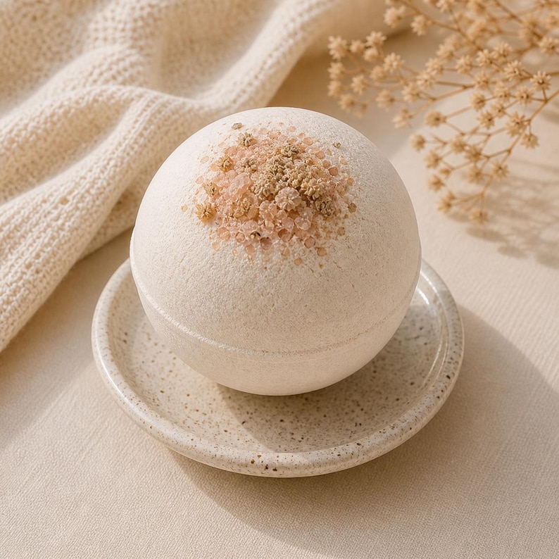 May include: A round, off-white bath bomb with a textured surface, topped with a cluster of light brown and pink crystals. It rests on a small, speckled ceramic dish. The background includes a cream-colored knitted blanket and dried flowers.