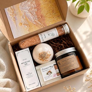 May include: A gift box filled with self-care items. Includes a postcard with an abstract beach scene, bath salts, a bath bomb, multi-use oil, coconut milk soap, and a scented candle. The box is brown and filled with brown paper. The items are arranged on a white surface.