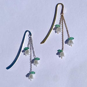 Puede incluir: Dos marcadores de metal, uno plateado y otro dorado, cada uno adornado con delicados dijes de flores blancas. Los dijes están suspendidos de cadenas y presentan pequeñas hojas verdes, creando un elegante diseño floral. Los marcadores están sobre un fondo blanco.