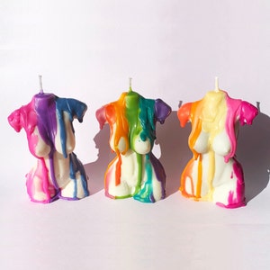 Op de afbeelding: Drie kleurrijke kaarsen in de vorm van een torso, elk met een ander kleurenschema: paars, roze en blauw; oranje, geel en groen; en geel, wit en roze.