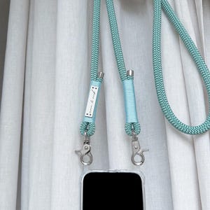 Pode incluir: Uma alça de telefone azul claro com fechos prateados e um pequeno rótulo branco que diz "Love & Rebel". A alça está presa a uma capa de telefone transparente com uma tela preta.