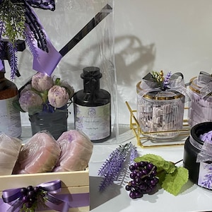 May include: A lavender-themed gift set with a clear plastic bag containing a lavender spray bottle, a dark glass bottle of lavender extract, and a small bouquet of lavender flowers. The set also includes a wooden box with three lavender-scented soaps, two glass jars with lavender-scented bath salts, and a black jar with a lavender-scented candle.